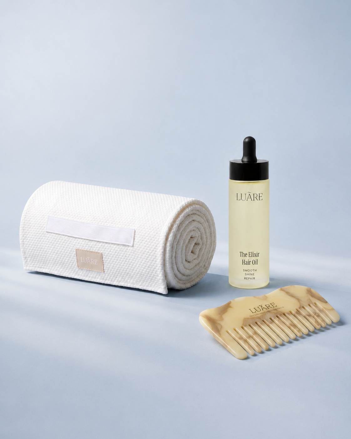 A Luâre hair care set on a light blue background, featuring a rolled white microfibre towel, a clear bottle of hair oil with a black dropper cap, and a hand-made plant cellulose comb.